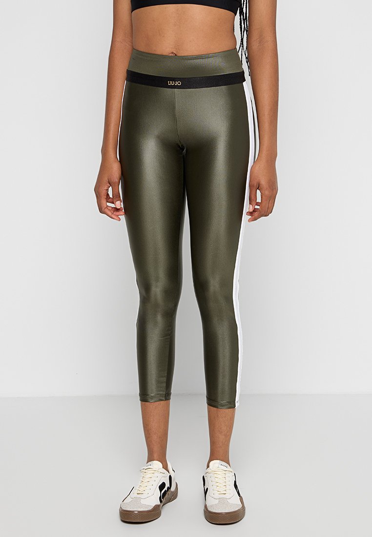 Mujer vistiendo leggins brillantes de color verde oliva con rayas laterales blancas y cintura negra, combinados con zapatillas blancas y negras.