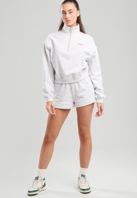 Vaaleanharmaa vetoketjullinen collegepaita, jossa on korkea kaulus ja resorireunat, yhdistettynä samanvärisiin shortseihin. Korostettu punaisella brodeeratulla tekstillä.