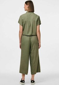 Combinaison verte olive à manches courtes, coupe décontractée, taille élastique, jambes larges, détail de pli au dos, portée avec des baskets noires.
