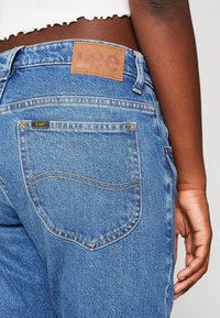 Des jeans en denim bleu présentent une patch en cuir marron avec le logo "Lee" à la taille, et des coutures visibles sur la poche arrière.