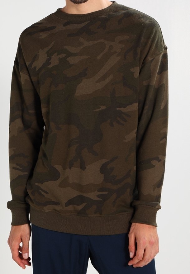 Homme portant un sweat-shirt à manches longues marron et vert foncé à motif camouflage et un pantalon foncé sur un fond uni.