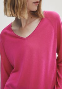 Pull en V fuchsia à manches longues, texture lisse et épaules raglan, avec un col net et une profondeur de couleur subtile.