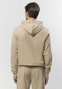 Bee¾värvi kapuutsiga pusa tekstuurse pinnaga, mis on lõdva lõike ja pikkade varrukatega. Kapuuts on pehmelt vormitud ja lamedalt laotatud.