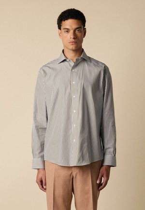 Homme portant une chemise à rayures verticales à manches longues et un pantalon marron clair, debout devant un fond beige uni.