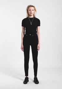 Młoda kobieta o blond włosach, ubrana w czarny t-shirt, czarne spodnie z wysokim stanem, czarne trampki i srebrne naszyjniki, stojąca na białym tle.