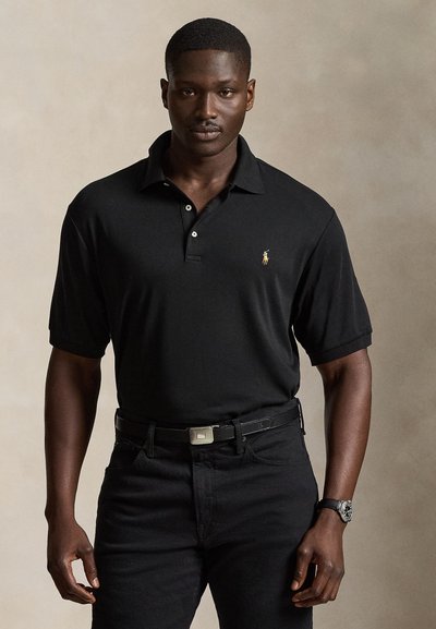 Mann trägt ein schwarzes kurzärmeliges Polohemd und schwarze Hose mit Gürtel, steht vor einem schlichten neutralen Hintergrund.