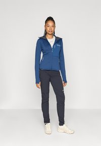 Casaco de fleece azul com fecho éclair e capuz cinza, apresentando dois bolsos frontais; combinado com calças azul marinho e sapatilhas de cor clara.
