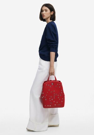 Red leather backpack with silver grommet accents, featuring double handles and a structured shape, paired with white wide-leg pants and a navy sweatshirt.