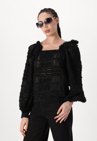 Schwarzes Cropped Top aus strukturiertem Stoff, mit durchsichtigen Lagen und puffärmel. Kombiniert mit schwarzen Hosen und übergroßen dunklen Sonnenbrillen.