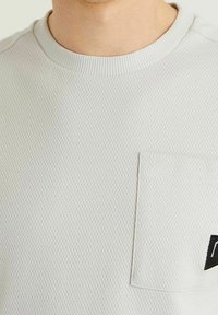 Jasnoszara, teksturowana bluza z krótkim rękawem, z ryflowanym kołnierzem oraz kieszenią na piersi z czarnym akcentem logo.