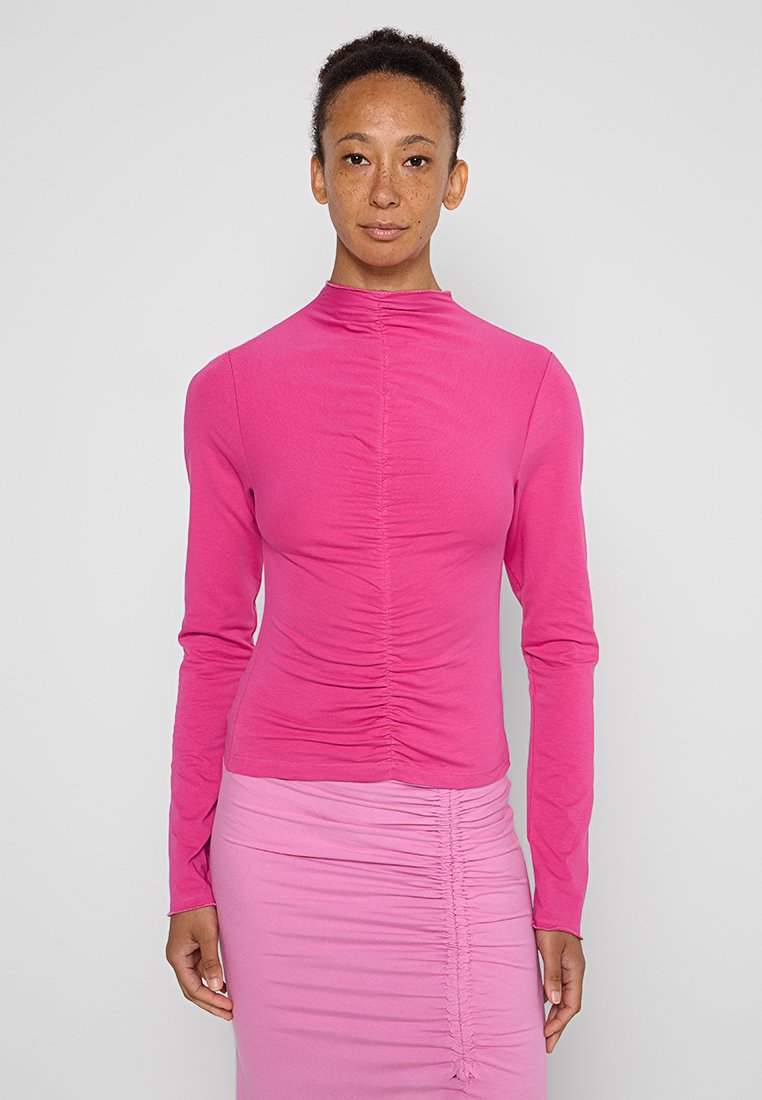 Langärmliges, eng anliegendes Oberteil aus kräftigem, einfarbigen pinken Baumwollstoff, mit Raffung an der Vorder- und Rückseite sowie einem hohen Kragen.