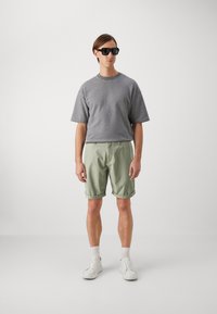 Felpa grigia a maniche corte, pantaloni corti arrotolati verde chiaro, sneakers bianche e occhiali da sole neri indossati da una figura maschile in piedi contro uno sfondo neutro.
