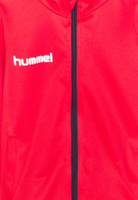 Röd hoodie med dragkedja framtill, tillverkad av mjukt tyg, med en marinblå dragkedja och en vit "hummel"-logotyp på vänster bröst.