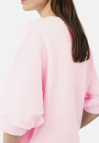 Helles rosa Sweatshirt mit Rundhalsausschnitt, kurzen puffigen Ärmeln und einer weichen Textur. Nahtdetails sind an den Ärmeln und am Rücken sichtbar.