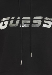 Černá mikina s kapucí, která se vyznačuje výrazným stříbrným logem "GUESS" na přední straně, má šňůrky na stažení a kovové koncovky. Viditelná je jemná textury látky.