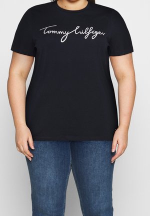 Person iført sort T-shirt med korte ærmer med hvid "Tommy Hilfiger"-tekst på brystet og blå denim jeans foran en ensfarvet baggrund.