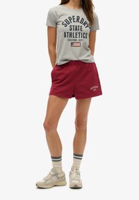 Camiseta de algodón gris con estampado "Superdry State Athletics", pantalones cortos de algodón burdeos y zapatillas ligeras con acentos azules y grises.