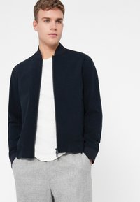 Námořnická bomber bunda s předním zipem, pružnými manžetami a kapsami, nošena přes bílou košili. Model stojí se založenýma rukama v kapsách.