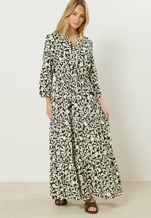 Kvinde iført en sort og hvid blomsterlangærmet maxikjole med indsnævret talje og lagdelt skørt, kombineret med brune sandaler.