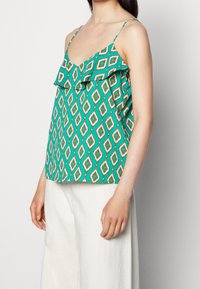 Haut sans manches teal avec un motif en losange et des détails en volants porté avec un pantalon large blanc taille haute, présenté des épaules aux hanches.