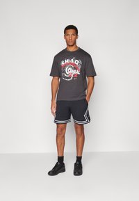 Sort grafisk T-shirt med rød og hvid design, parret med sorte sportsshorts med hvide striber og sorte sneakers.