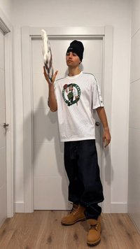 Ung man inomhus klädd i Boston Celtics-tröja, svart mössa, mörka byxor och bruna stövlar, kastar upp ett vitt och grått föremål i luften.