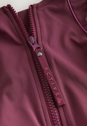 Lähivõte marooni värvi tõmblukukeelel, millel on sõna "ACTIVE" ja kolmnurkse logoga tõmmitsal.