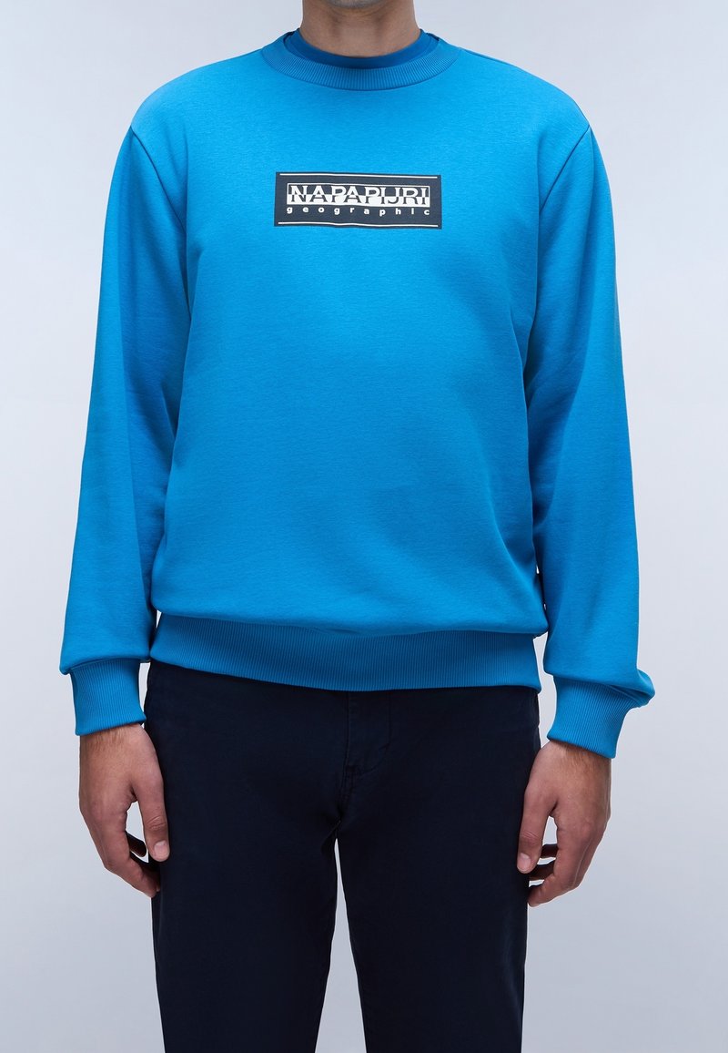 Homme portant un sweat-shirt bleu vif à manches longues avec un logo rectangulaire noir et blanc indiquant "NAPAPIJRI geographic" sur la poitrine.