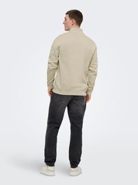 Homme portant un sweat-shirt beige, un jean noir et des baskets blanches, debout de dos devant un fond uni gris clair.