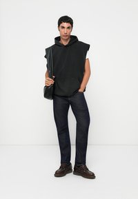 Schwarze ärmellose Kapuzenjacke mit einer Fronttasche, kombiniert mit dunkelblauen Jeans und braunen Schnürschuhen, Tasche über der Schulter vor einem schlichten Hintergrund.