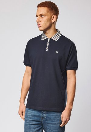 Młody mężczyzna o krótkich kręconych włosach, ubrany w granatową koszulkę polo z kraciastym kołnierzykiem i jasnoniebieskie dżinsy, spoglądający na bok.