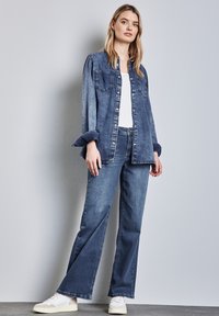Giacca di jeans blu a due tonalità, con bottoni e tasche sul petto. Abbinata a jeans a gamba larga blu chiaro e sneakers bianche.