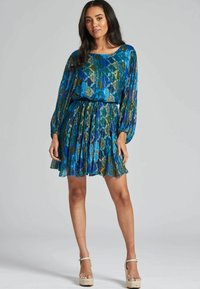 Vestido azul y verde con un patrón, de mangas largas y cintura ajustada. Falda acampanada con volantes, combinado con zapatos de plataforma de color claro.