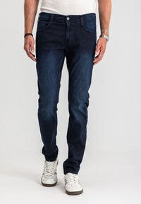 Mężczyzna ubrany w ciemnoniebieskie dżinsy, białe trampki, białą koszulkę, srebrny zegarek na lewej ręce oraz czarną bransoletkę na prawej, stojący.