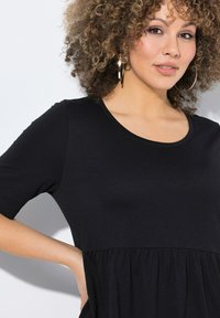 Černé šaty s krátkým rukávem z jemné látky, s kulatým výstřihem a nařasenou sukní, která dodává objem. Zlato-kroužkové náušnice dotvářejí vzhled.