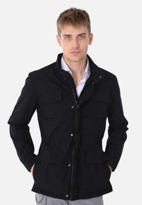 Giacca utility nera con chiusura a zip, quattro tasche frontali e colletto rialzato. Realizzata in un tessuto resistente, presenta una texture liscia.