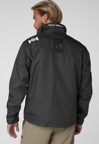 Chaqueta impermeable para hombre en gris oscuro, con cuello alto, acentos de logo y un panel de ventilación en la parte posterior. Presenta una textura suave y ligera.