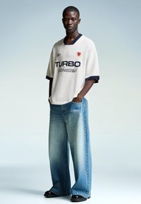 Oversized cream jersey with navy accents, “TURBO” in bold black text, paired with wide light blue denim jeans and black shoes.