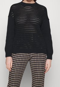 Svart stickad tröja med öppet mönster över kroppen, ribbade ärmslut, i kombination med svart och beige rutiga byxor.