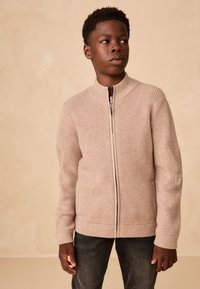 Pull à fermeture éclair en maille côtelée beige avec col montant et manches longues, avec un patch sur le bras gauche. Associé à un jean foncé.