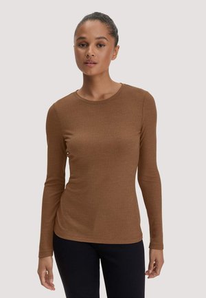Frau trägt ein braunes, geripptes Langarmshirt und schwarze Hose und steht vor einem schlichten hellen Hintergrund.