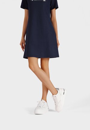 Abito navy a maniche corte con tessuto testurizzato e logo bianco. Il modello indossa scarpe da ginnastica bianche con accenti argentati e suola spessa.