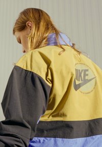 Nike-jakke i gul, svart og blå. Har en stor logo på baksiden, fargeblokkert design og slitesterkt stoff.