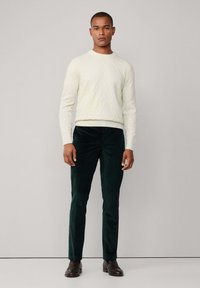Suéter de punto de cable color crema, pantalones de pana verde y zapatos marrón oscuro. El suéter tiene un escote redondo; los pantalones son ajustados con un corte recto.