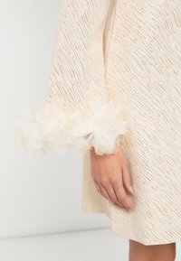 Rochie crem din material texturat, cu accente de flori plisate la manșete și design cu croială dreaptă.