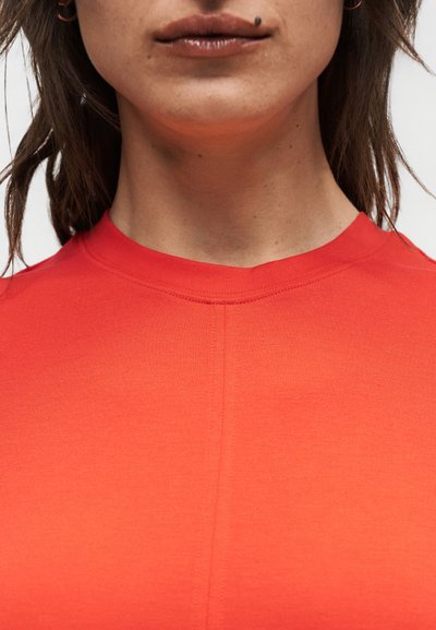 Κοντινό πλάνο του κάτω μέρους του προσώπου και του λαιμού ενός ατόμου που φοράει ένα φωτεινό κόκκινο μπλουζάκι με στρογγυλή λαιμόκοψη, με μια μικρή ελιά πάνω από το χείλος.