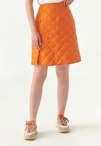 Falda acolchada naranja con cremallera diagonal, corte a la altura de la rodilla y una textura ligera. Combinada con zapatillas beige y naranjas.