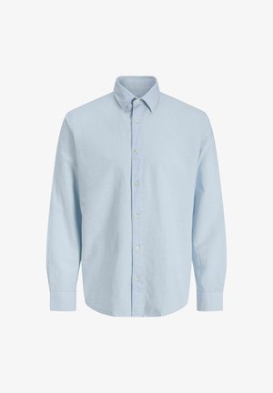 Ragazzo con camicia azzurro chiaro con bottoni, pantaloni blu scuro e scarpe da ginnastica bianche, in piedi, che guarda leggermente di lato su uno sfondo semplice.