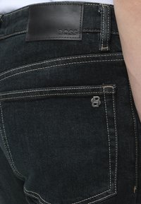 Gros plan de la poche arrière d'un jean en denim noir avec des coutures blanches, un petit logo métallique "B" et un patch en cuir noir avec "BOSS" embossé.