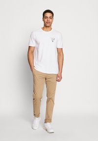 White short-sleeve t-shirt with a black graphic print on the chest, paired with beige chinos and white sneakers. Simple, casual design.